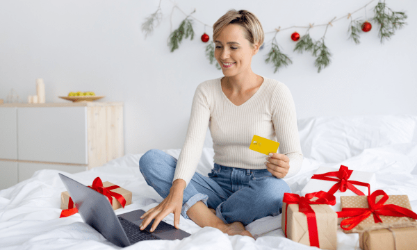 ragazza seduta sul letto, ha davanti un computer portatile e ha in mano una carta di credito, pronta per lo shopping natalizio